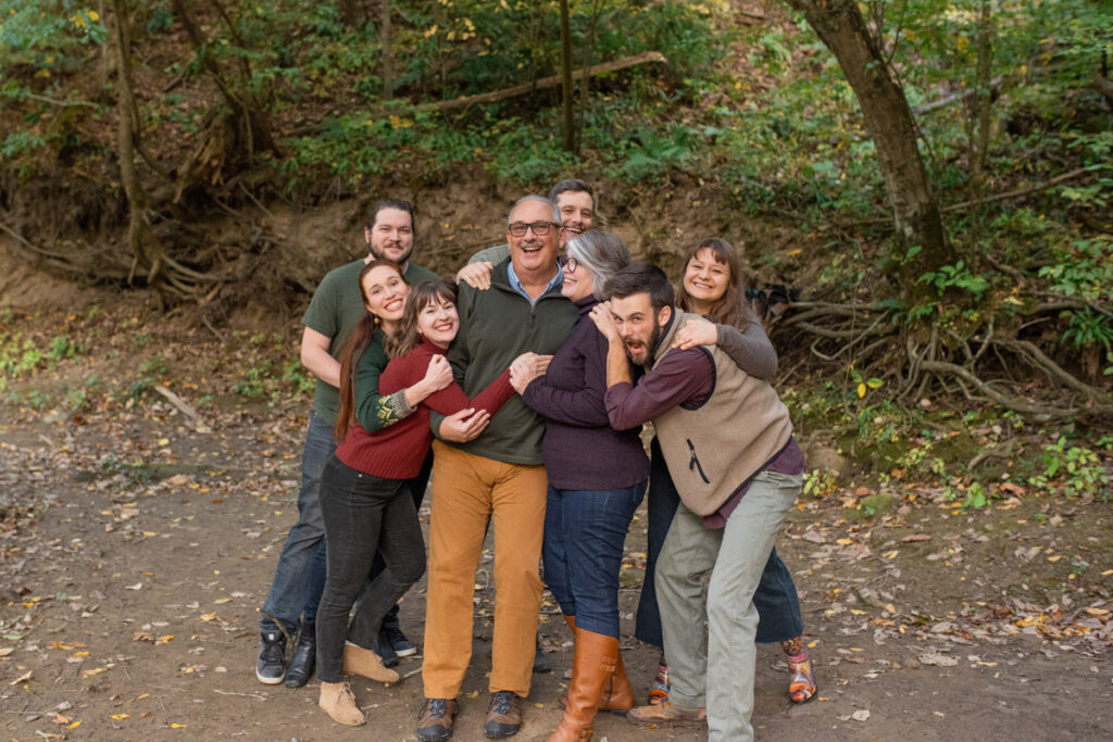 Adult children surprise hug their parents in a wooded area.