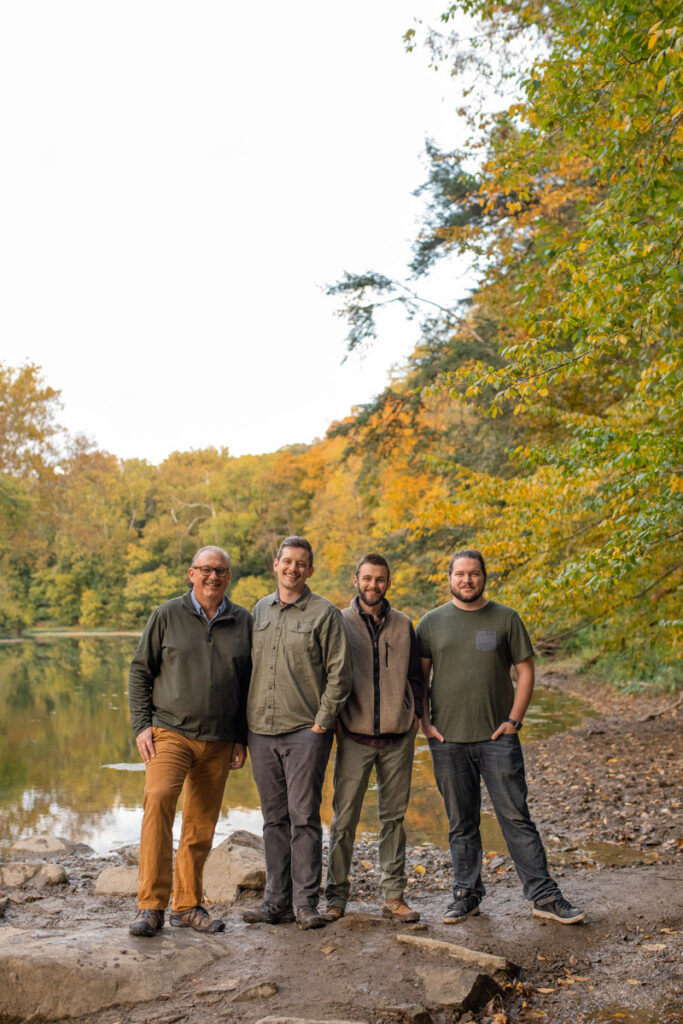 Four men stand together smiling next to creek in the fall.