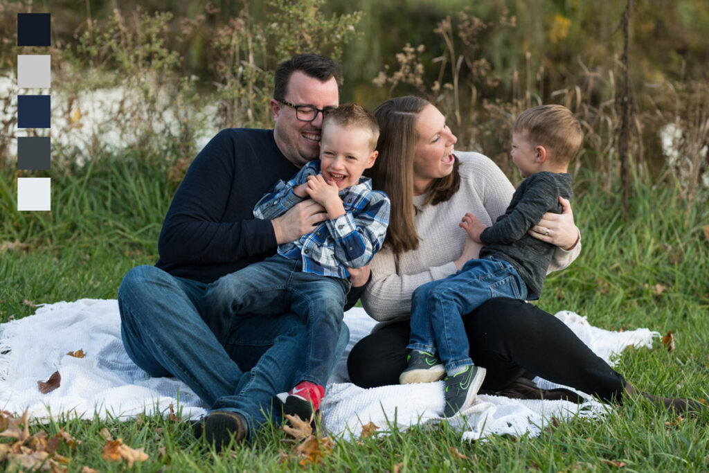 Parents tickle two sons while sitting on a white blanket near a pond.