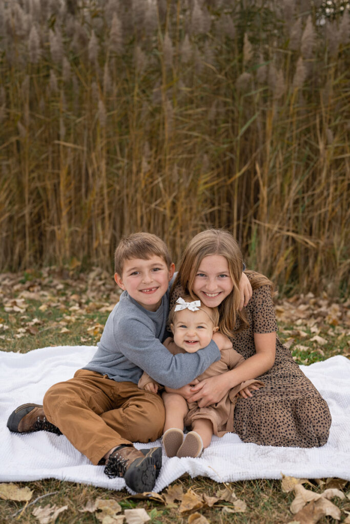 Three siblings sit on blanket with big smiles.