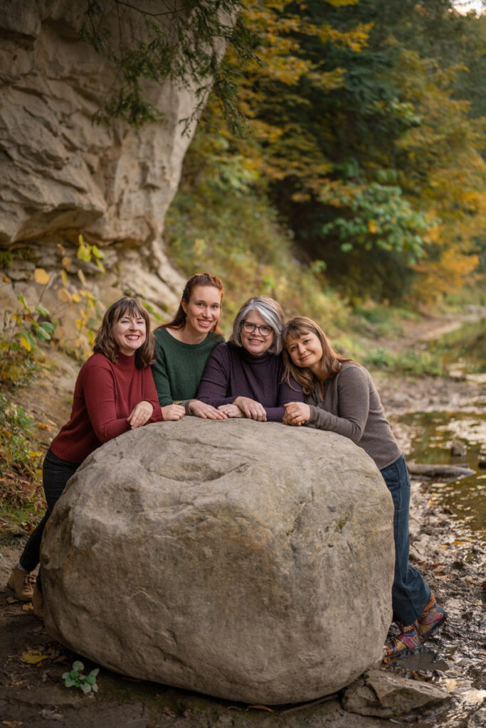 Four women lean against giant boulder near creek and smile.