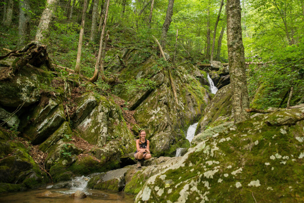 Woman crouches down on rock near waterfall.