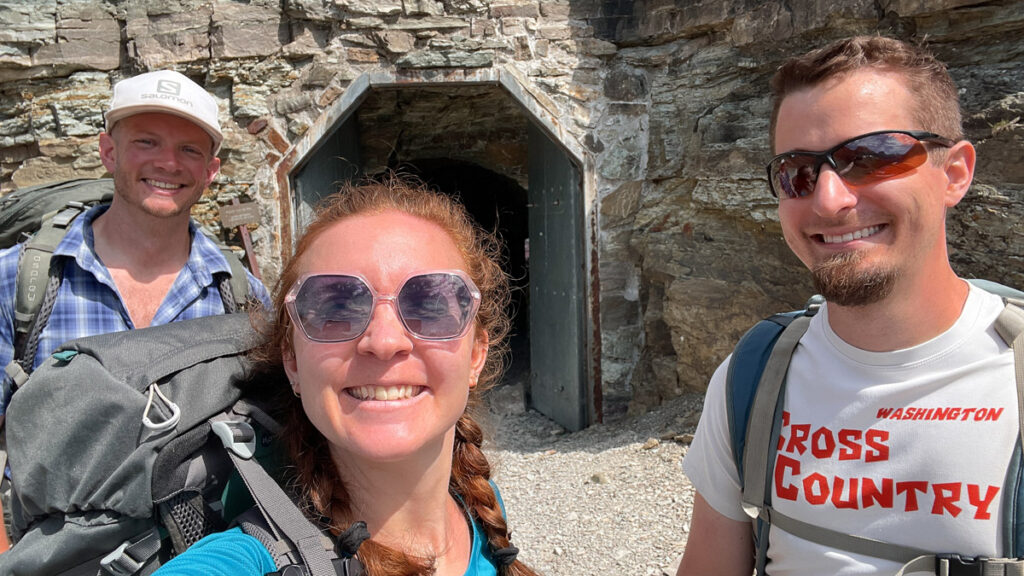 Three hikers smile with their packs before walking through mountain tunnel.