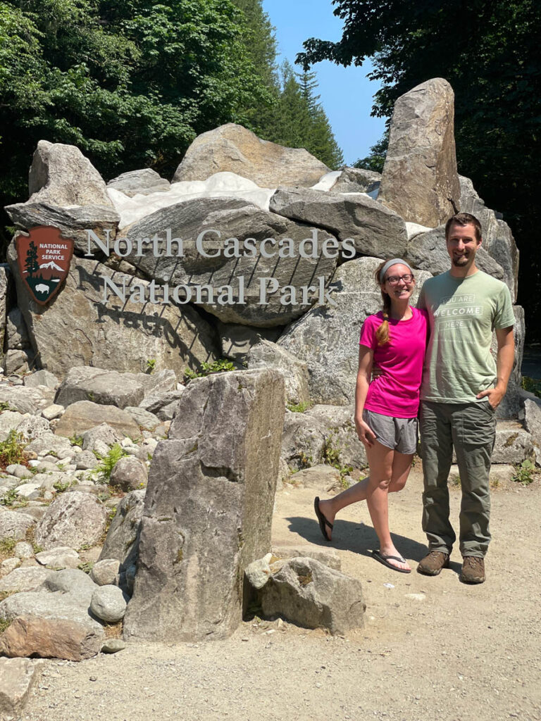 Couple stands in front of North Cascades National Park Sign.
