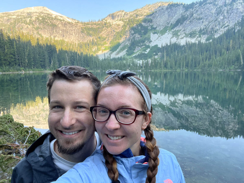 Hiking couple smile in front of lake with mountain reflection.