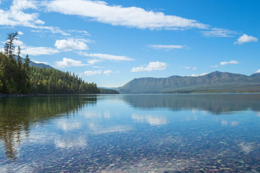 Blue skies and clouds reflect in lake at Glacier National Park with colorful rocks.