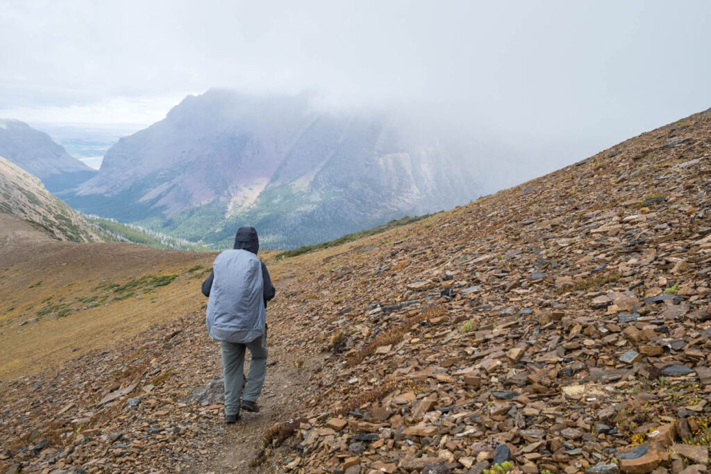Man is backpacking on a thin, rocky trail on the side of a mountain at Glacier National Park.