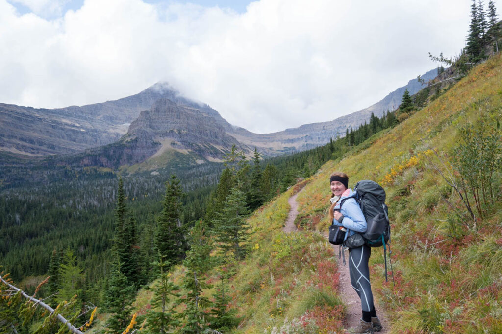 Woman is smiling on a small, dirt trail overlooking evergreen trees at Glacier National Park.