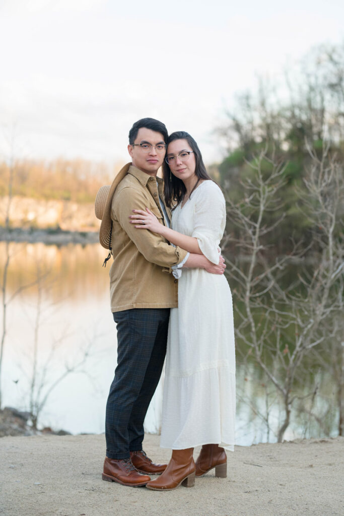 Couple embrace near water with serious expressions on their faces.