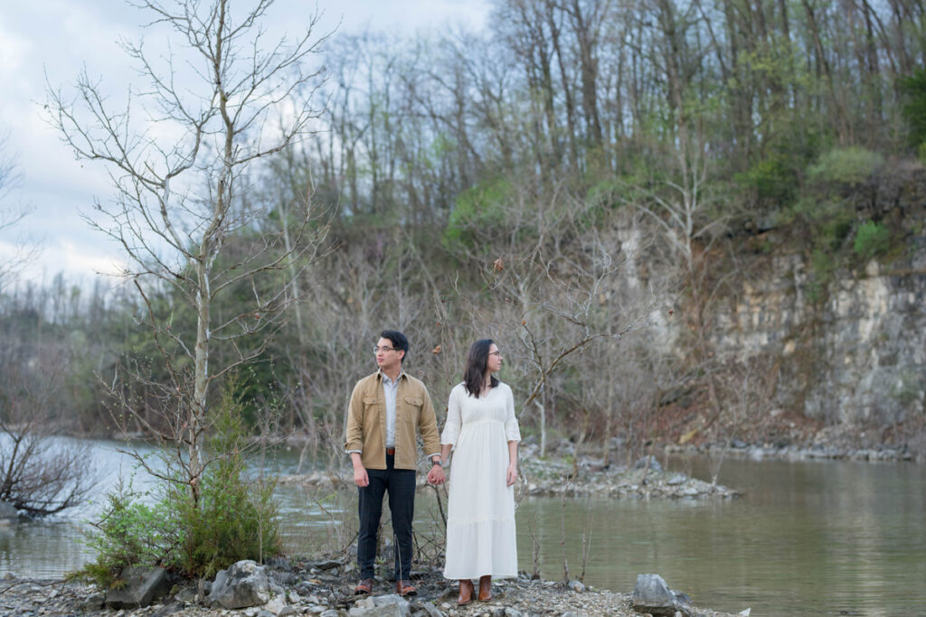 Couple holds hands while looking in opposite directions on rocky shore near lake.