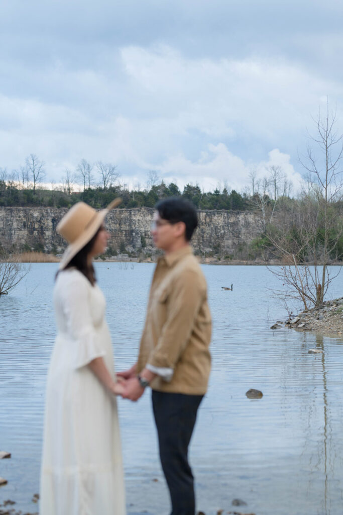 Couple holds hands near lake with geese and water behind them in focus.