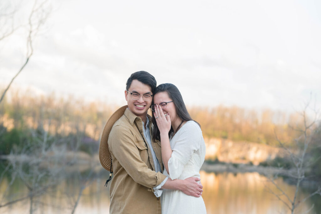 Couple laughs while hugging near water at sunset.