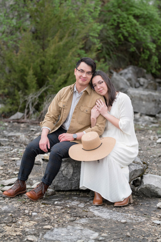 Man and woman site closely together on boulder and softly smile.