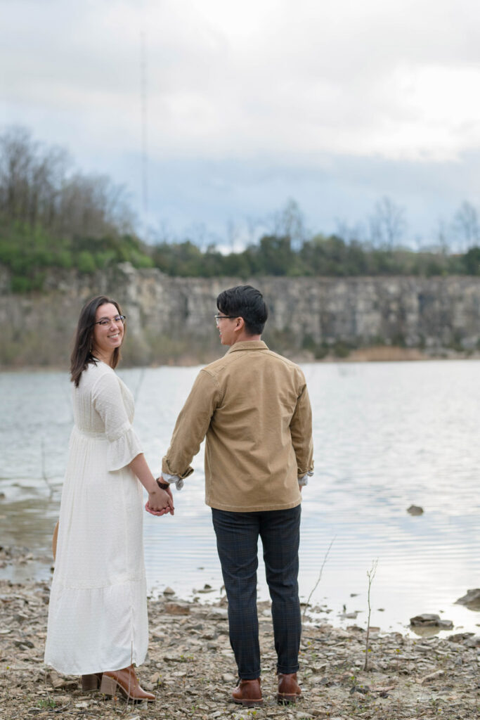 Man looks at woman while they walk together on rocky shore holding hands.