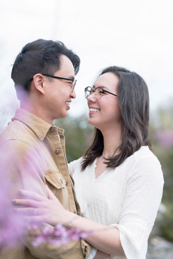 Couple smiles while looking at one another and standing among purple flowering tree.