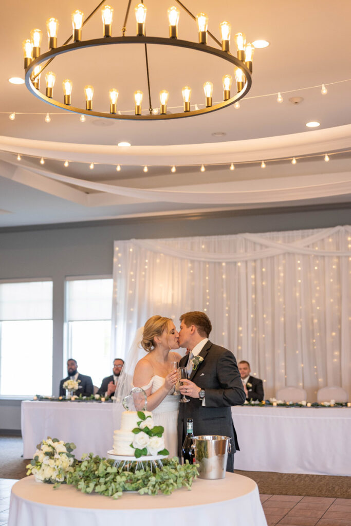Groom and bride share a kiss while holding champagne glasses at reception.