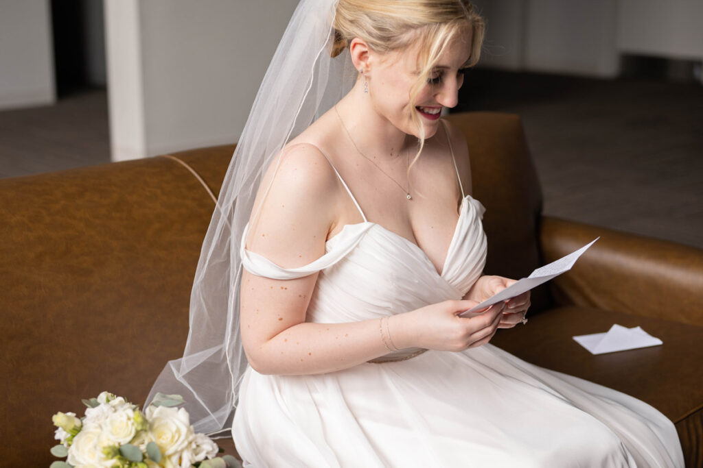 Bride reads letter from the groom on a tan leather couch.