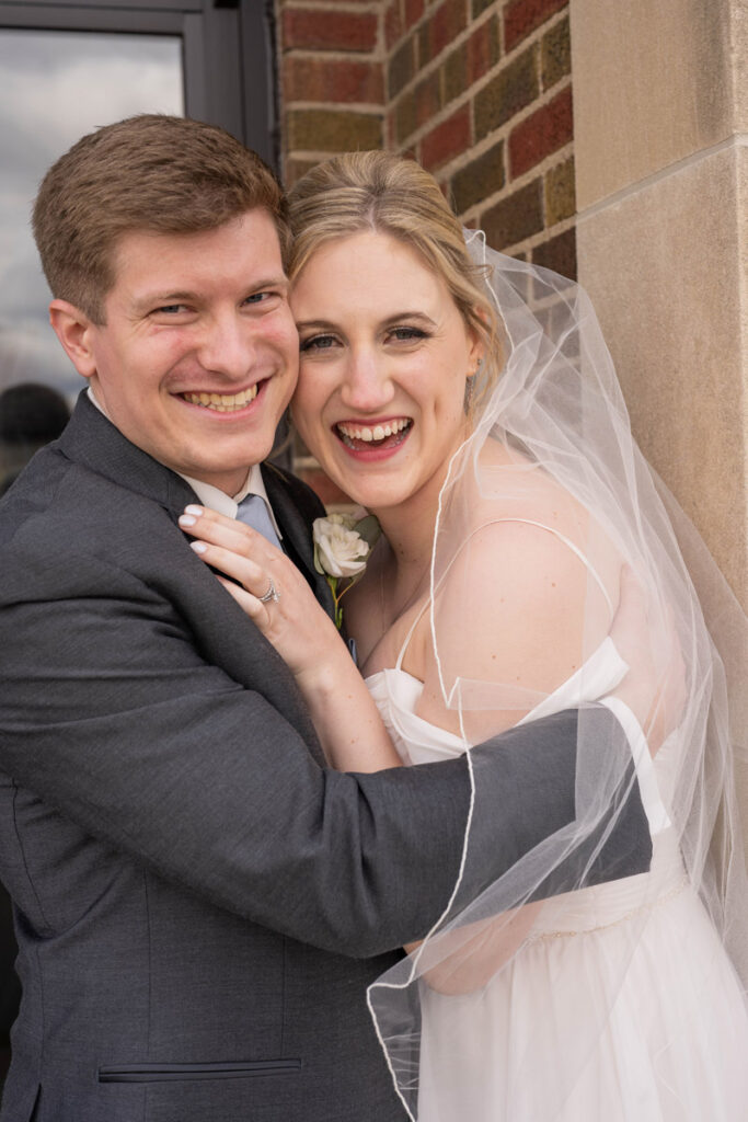Bride and groom holding each other close next to a brick wall and smiling happily.