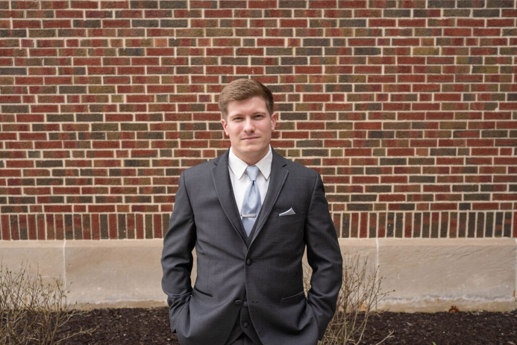 Groom with serious expression standing with hands in pockets in front of a brick wall.