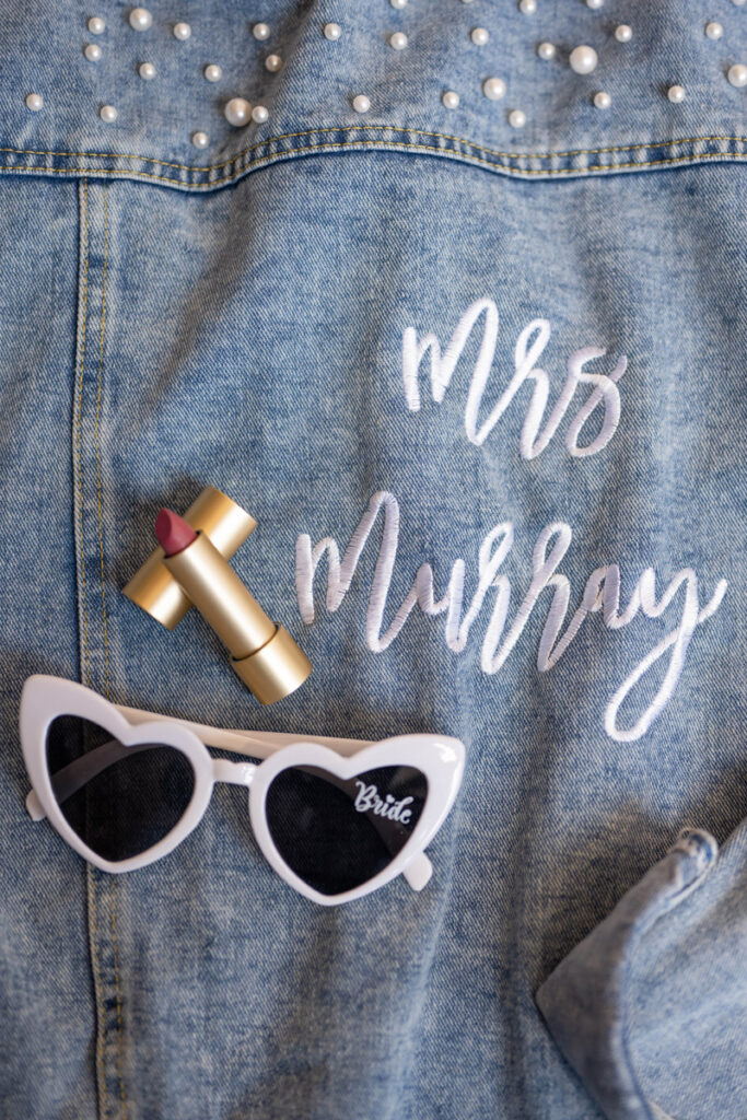Bride's jean jacket with her new name on it, custom wedding lipstick, and white heart sunglasses.