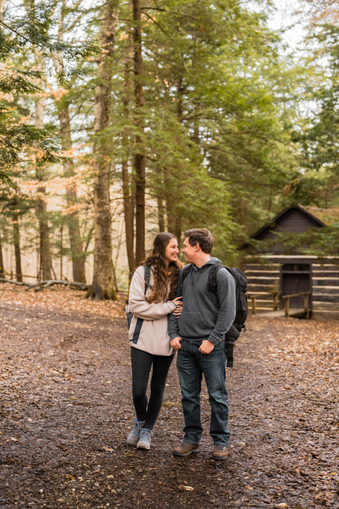 Hiking couple stands in front of log cabin with packs on.