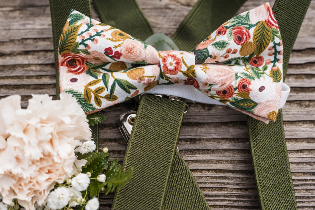 Floral bowtie sits on wooden table with green suspenders and bowtie.