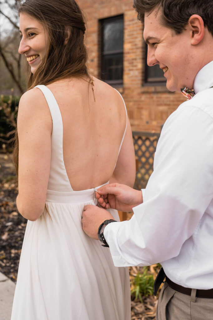 Groom and bride laugh while he zips up her dress.
