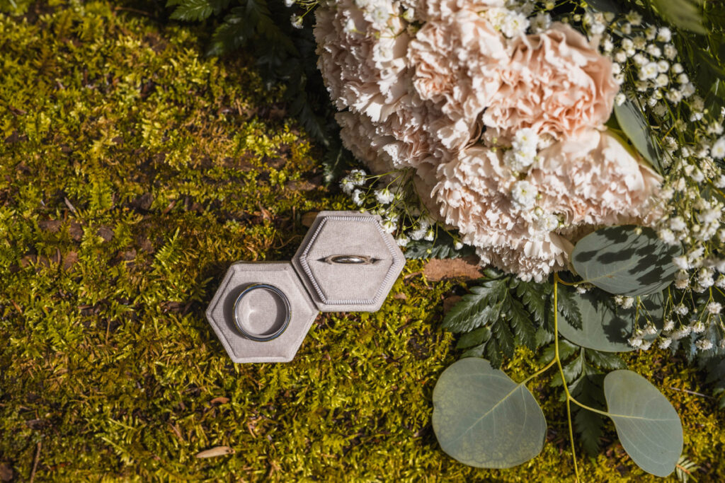 Wedding rings in a grey case resting on a mossy log next to pink carnations.
