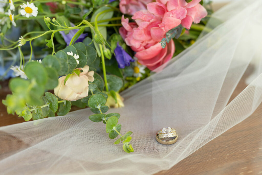 Gold wedding rings rest on a white veil with wildflowers behind them.