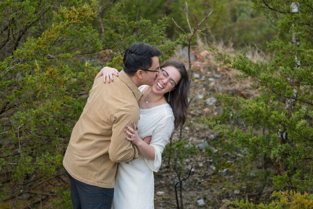 While standing in front of cedar trees, groom kisses bride while she laughs.