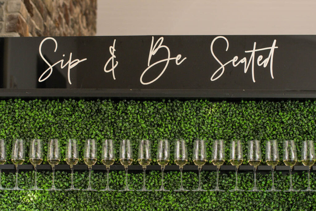 Champagne wall with greenery behind glasses and sign that reads Sip and Be Seated above.