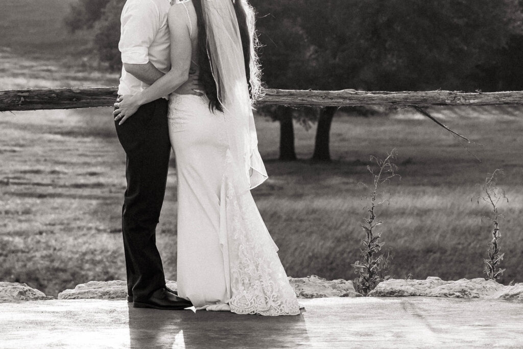 Couple embrace by wooden fence overlooking an open field.