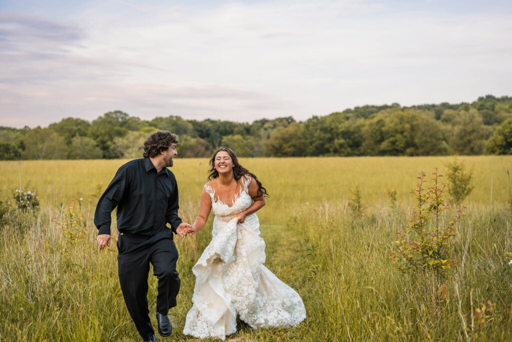 Bride and groom laughing while they run through a tall meadow.