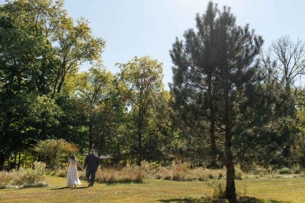 Bride and groom hold hands while walking through a meadow.