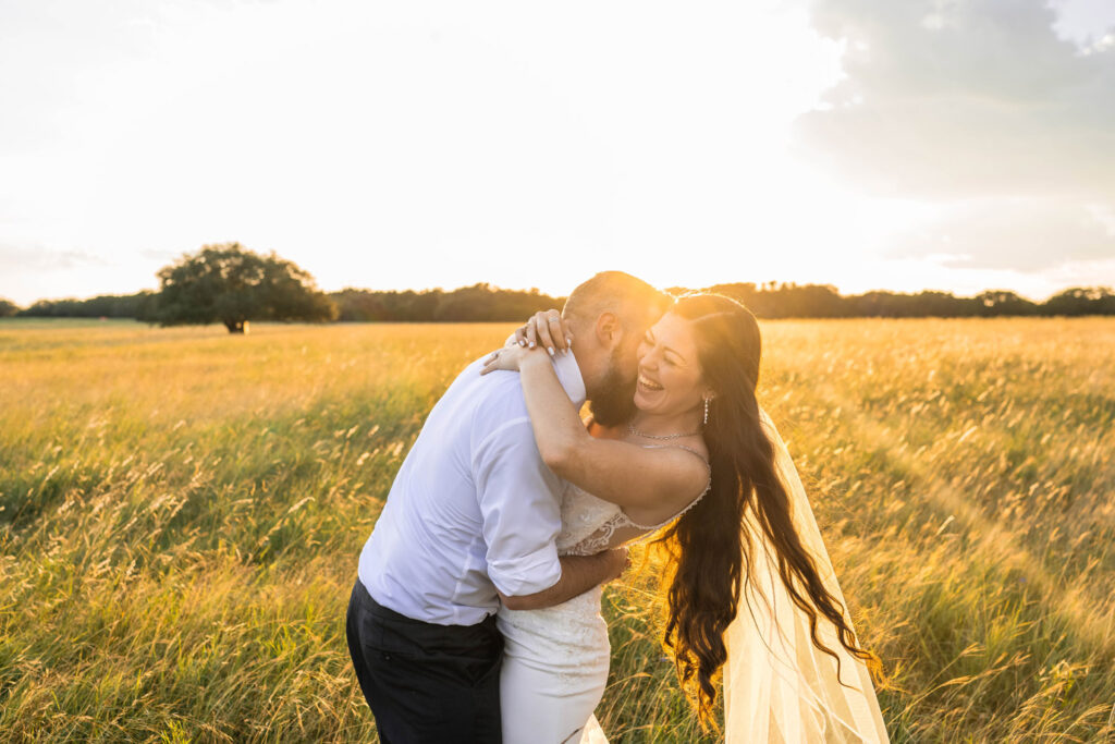 Groom kissing bride's neck making her laugh while sun sets behind them in an open field. 