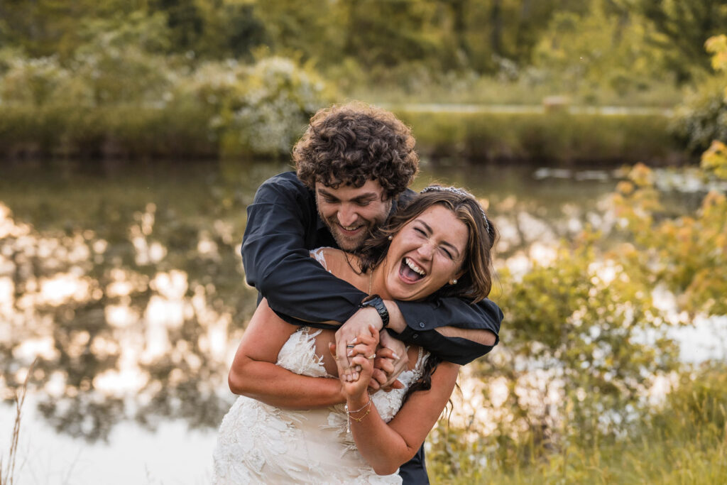 Bride and groom laughing and hugging one another while standing in front of a pond.