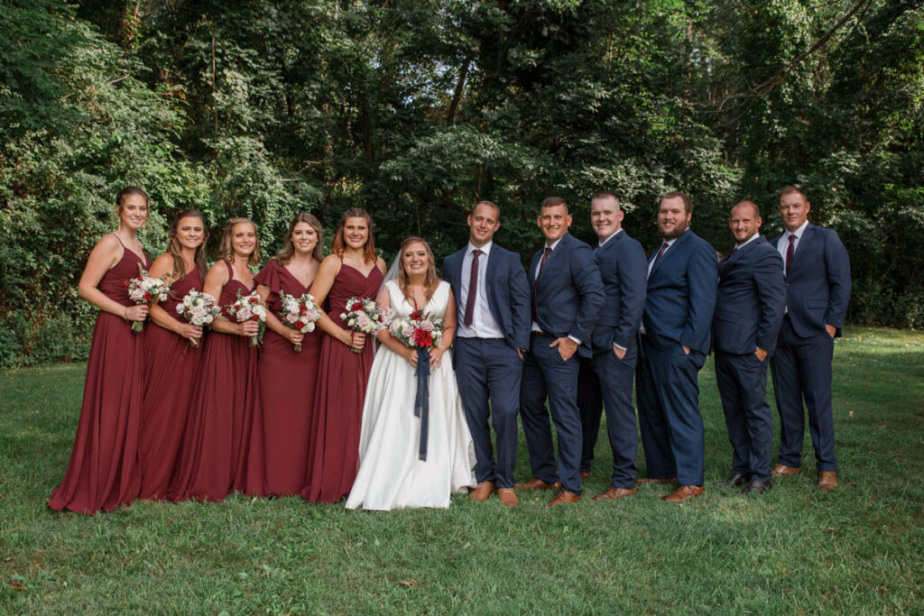 Bride and groom stand with their wedding party in front of a line of trees.