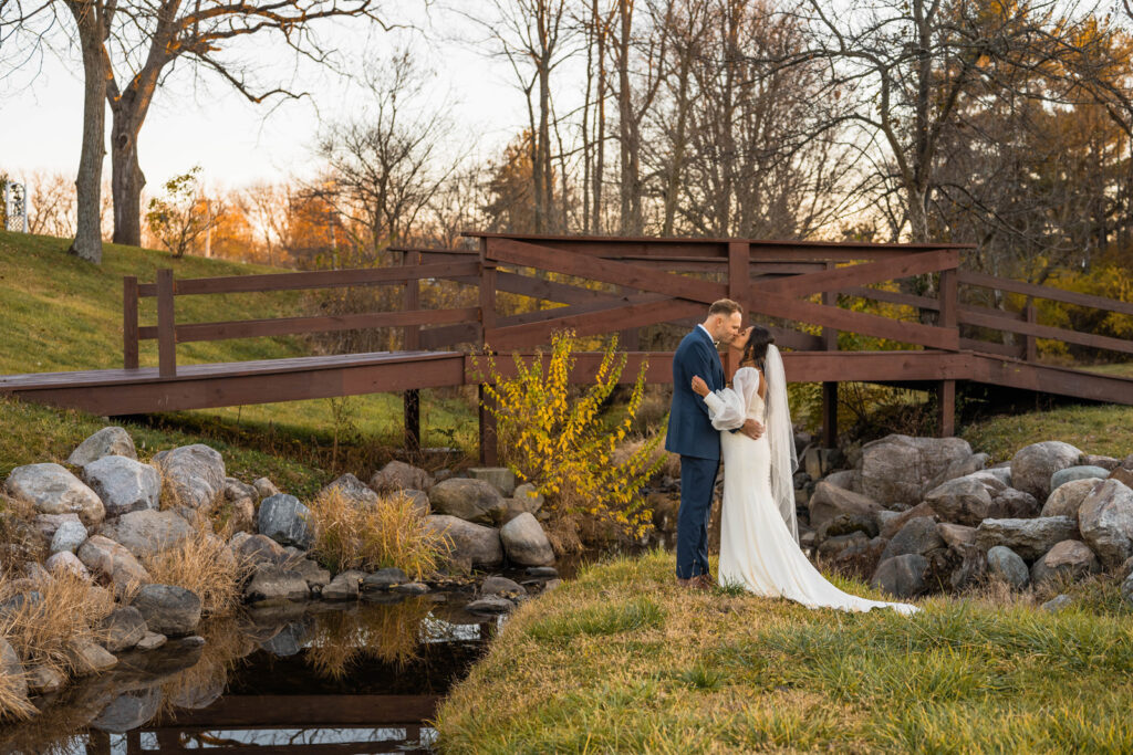 Couple kiss in front of bridge and small stream at sunset.