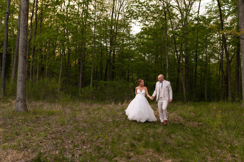 Bride and groom walk hand in hand next to forest at Owl Ridge Wedding and Event Center.