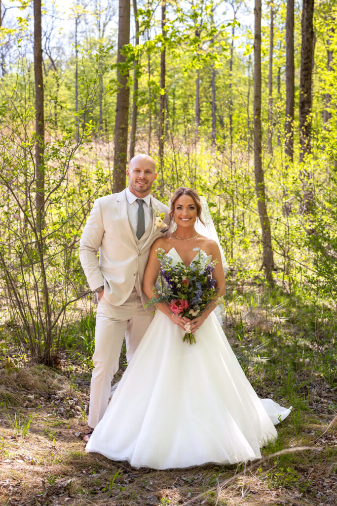 Bride and groom stand together smiling in front of a forest.