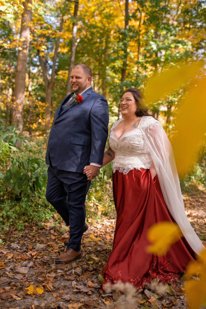 Bride and groom walk together among fall leaves holding hands.