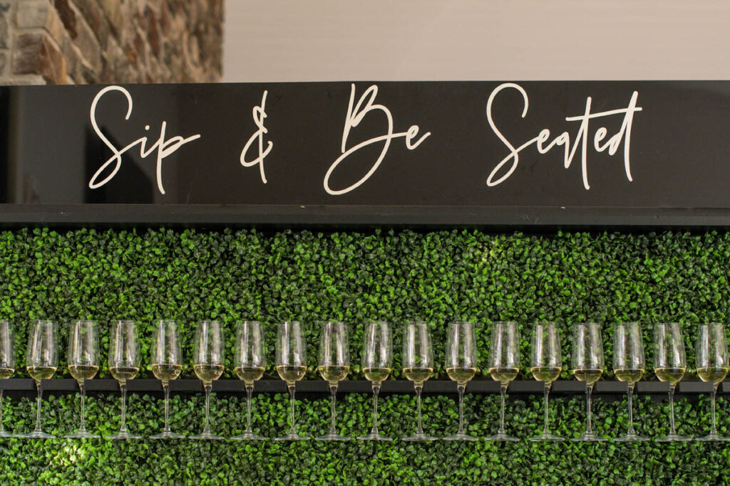 Sip and be seated champagne wall with greenery at Owl Ridge Wedding Venue.