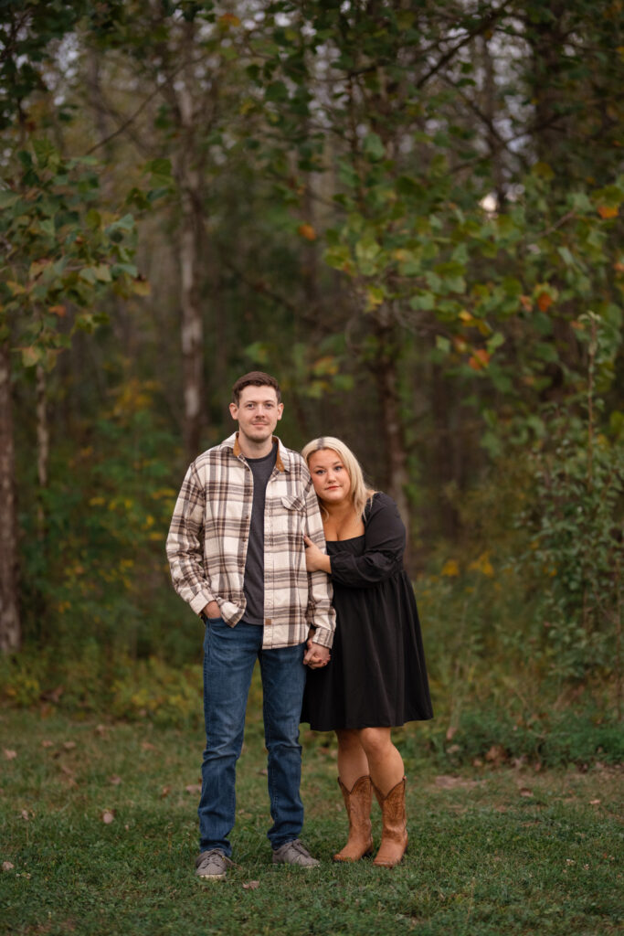 Engaged couple stands looking serious in front of wooded area at Washington Township Park.