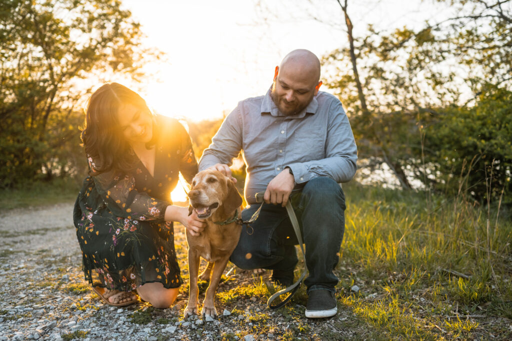 Engaged couple loves on their dog as the sun sets behind them.