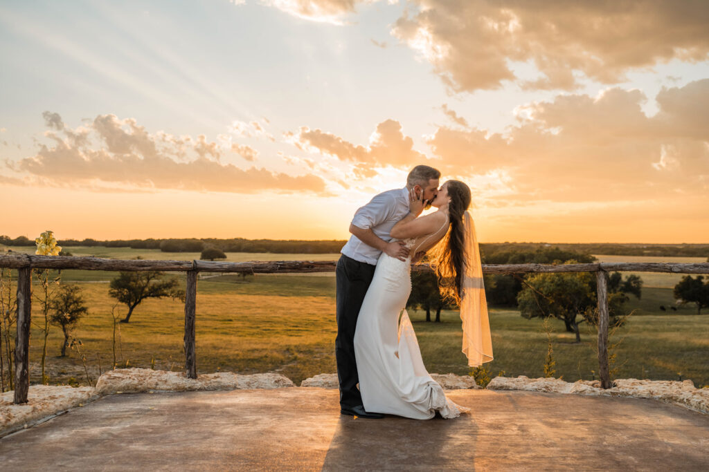 Bride and groom share a kiss at sunset overlooking a ranch.