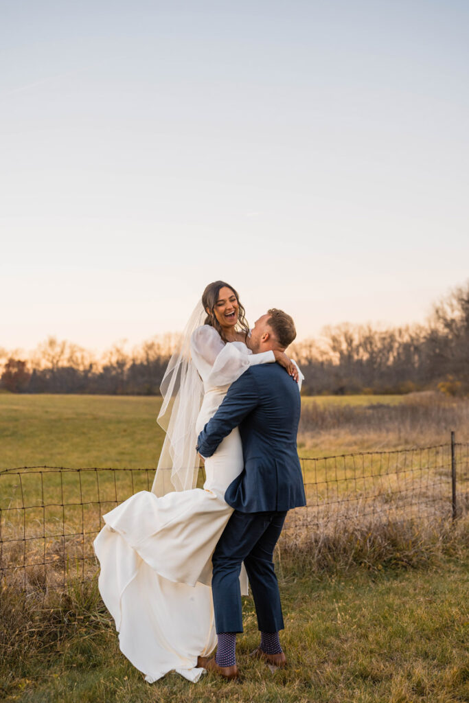 Groom picks up bride while she laughs at sunset.