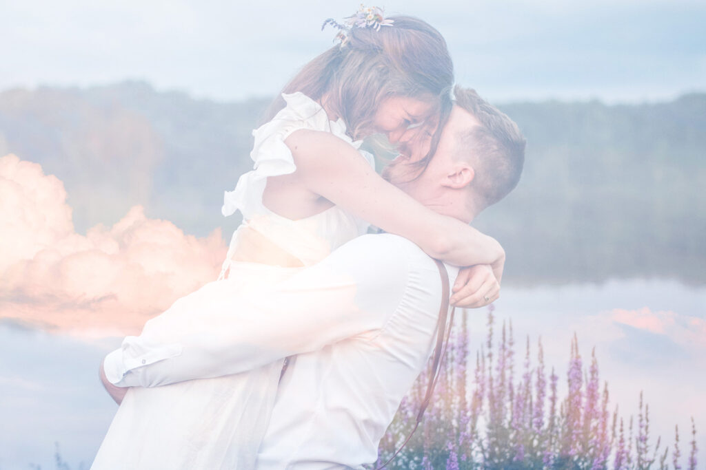 Groom picks up bride with a double exposure of the clouds at sunset.