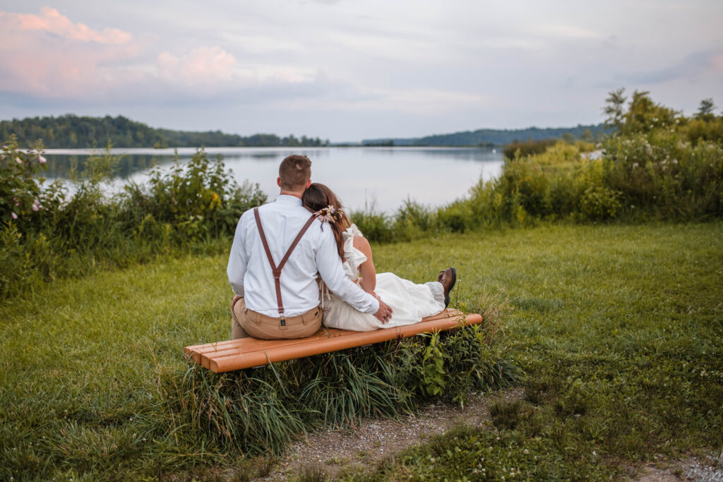 Groom and bride sit together watching the sun set over the water at Eagle Creek Park.