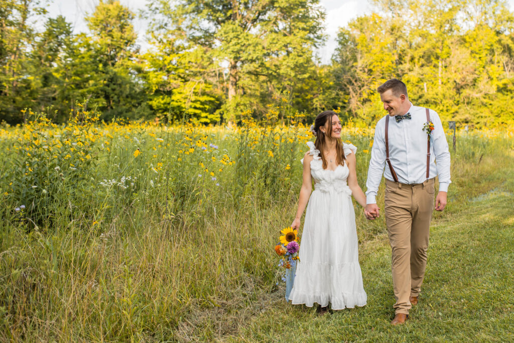 Couple holds hands during wildflower elopement as they walk next to a field of flowers.