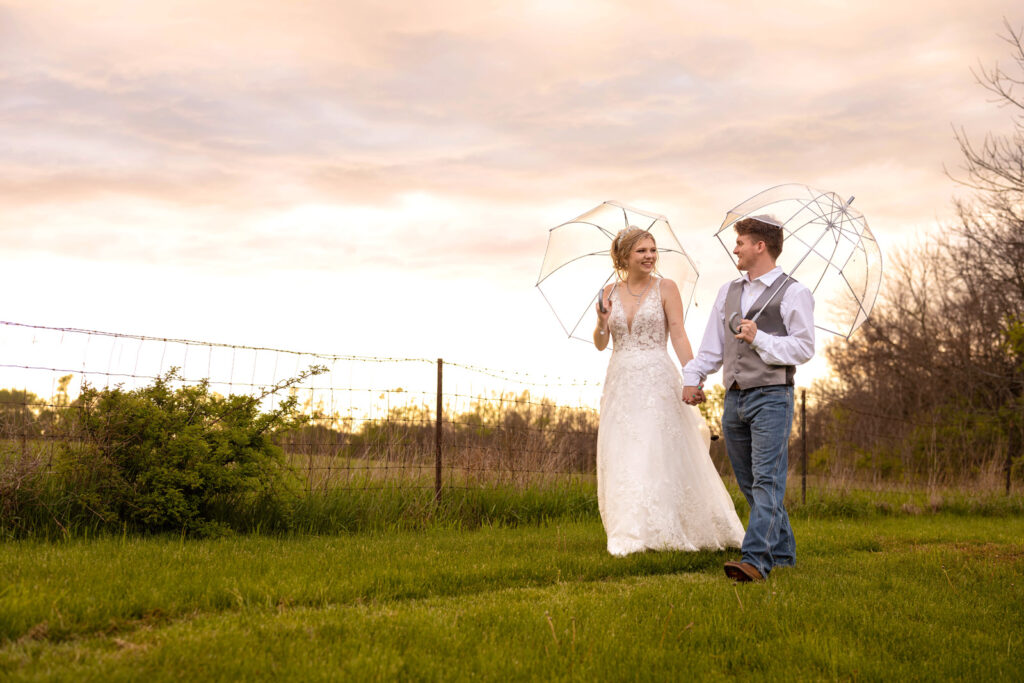 For their rainy, spring wedding day, a couple walks through the grass at Ash and Oak Weddings holding umbrellas.